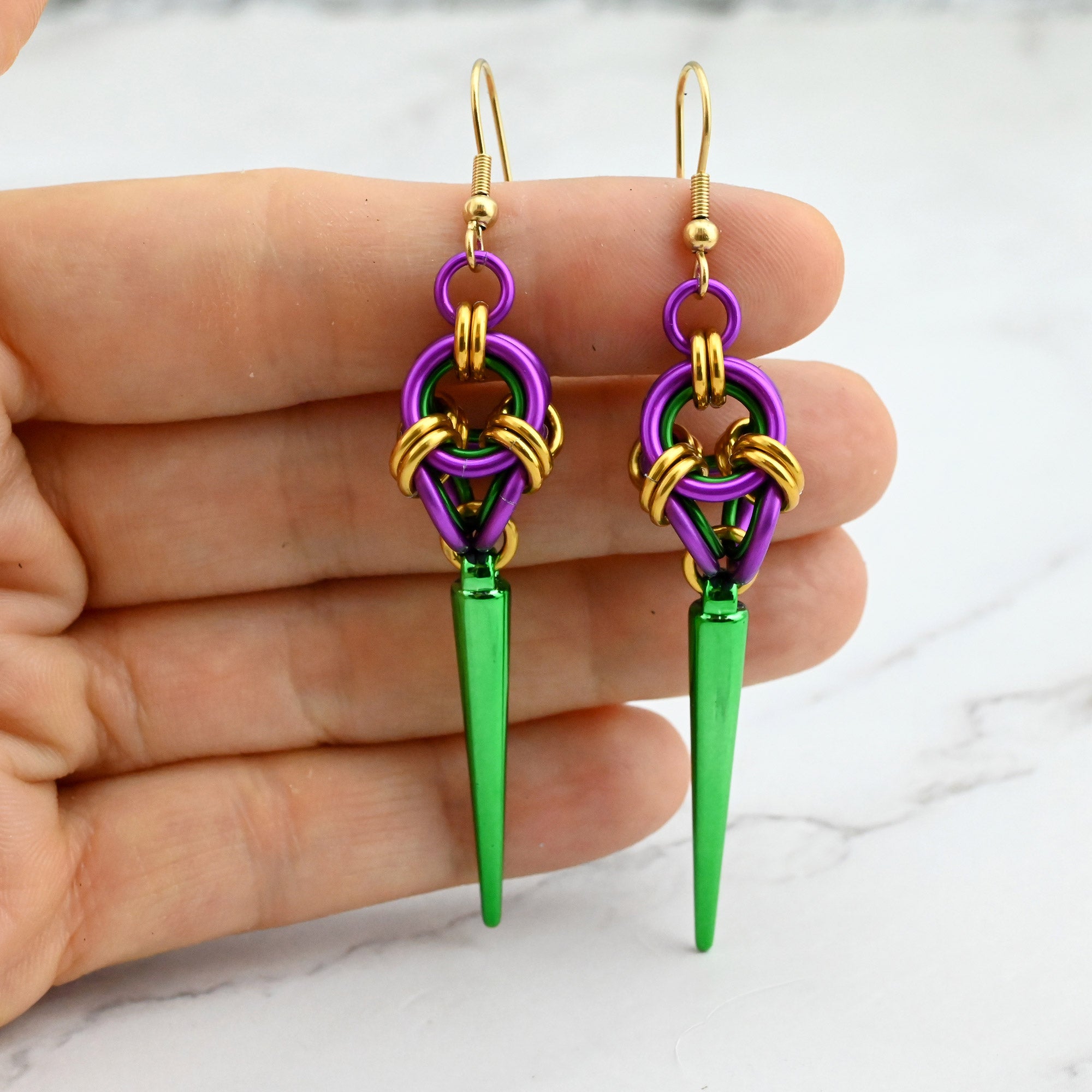 A hand holding the purple, gold, and green spiked polyhedron earrings to demonstrate their scale and size against fingers.