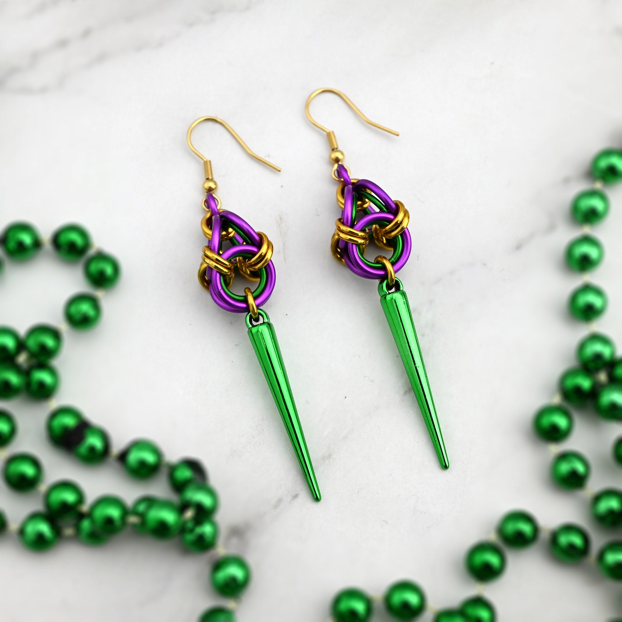 Top-down flat lay of Mardi Gras spiked earrings on a white marble surface surrounded by green metallic parade beads.