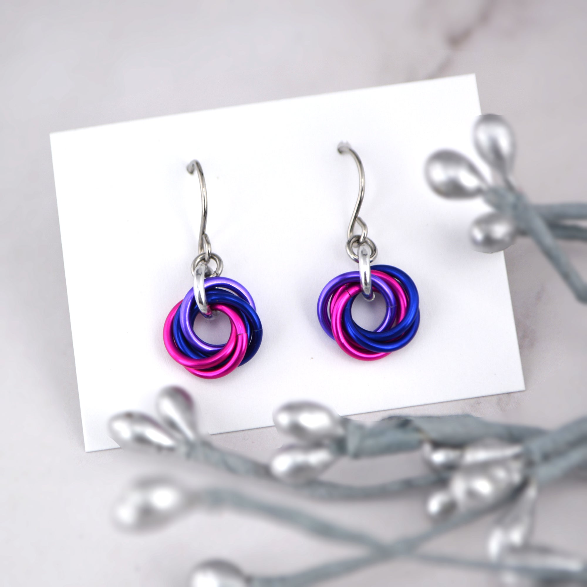 A pair of chainmaille Bisexual Pride Mini Knot Earrings are displayed on a small white card. The earrings feature interwoven links in the bisexual flag colors: pink, purple, and blue. Silver decorative elements are blurred in the foreground.