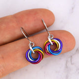 A close-up shot of a pair of polyamorous pride mini knot earrings, in colors to match the new polyamorous trim-color flag - blue, purple, yellow, pink, white. The earrings are rresting on chainmaille artisan Rebeca Mojica’s fingertips. The minimalist stainless steel ear wires are visible.