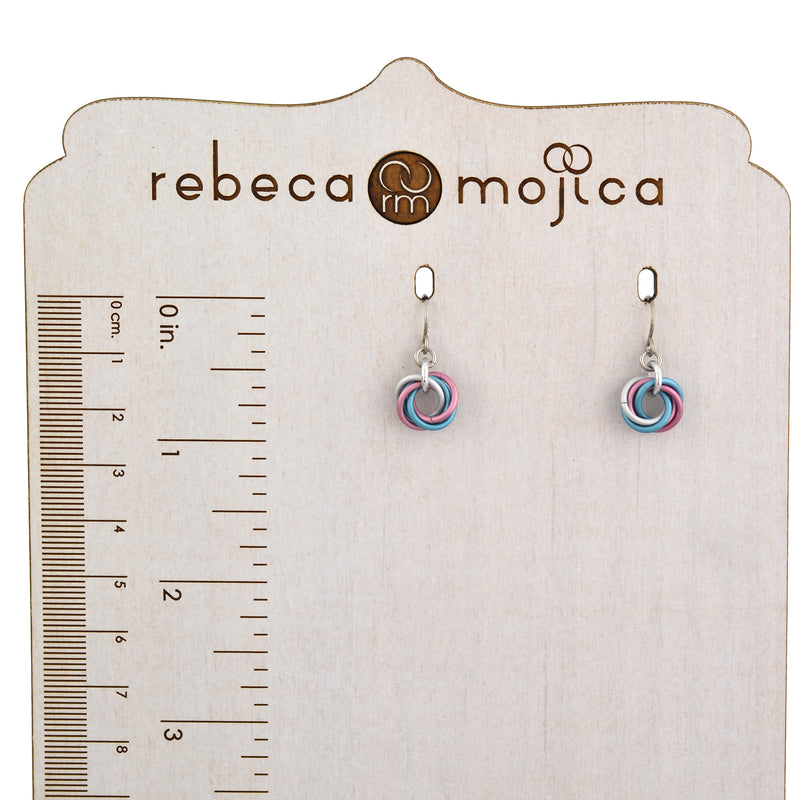 A pair of Transgender (light) flag mini knot earrings is displayed on a light wooden jewelry stand with a ruler printed on it, showing the earring's approximate 1-inch length. The "rebeca mojica" logo is at the top of the stand.