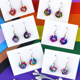 A flat lay photo showing six pairs of LGBTQ pride chainmaille mini knot earrings, each on a small white card, arranged on a rainbow pride flag background. The earrings shown include Genderfluid, Rainbow, Pansexual, Bisexual, Transgender (light), and Lesbian flag designs.