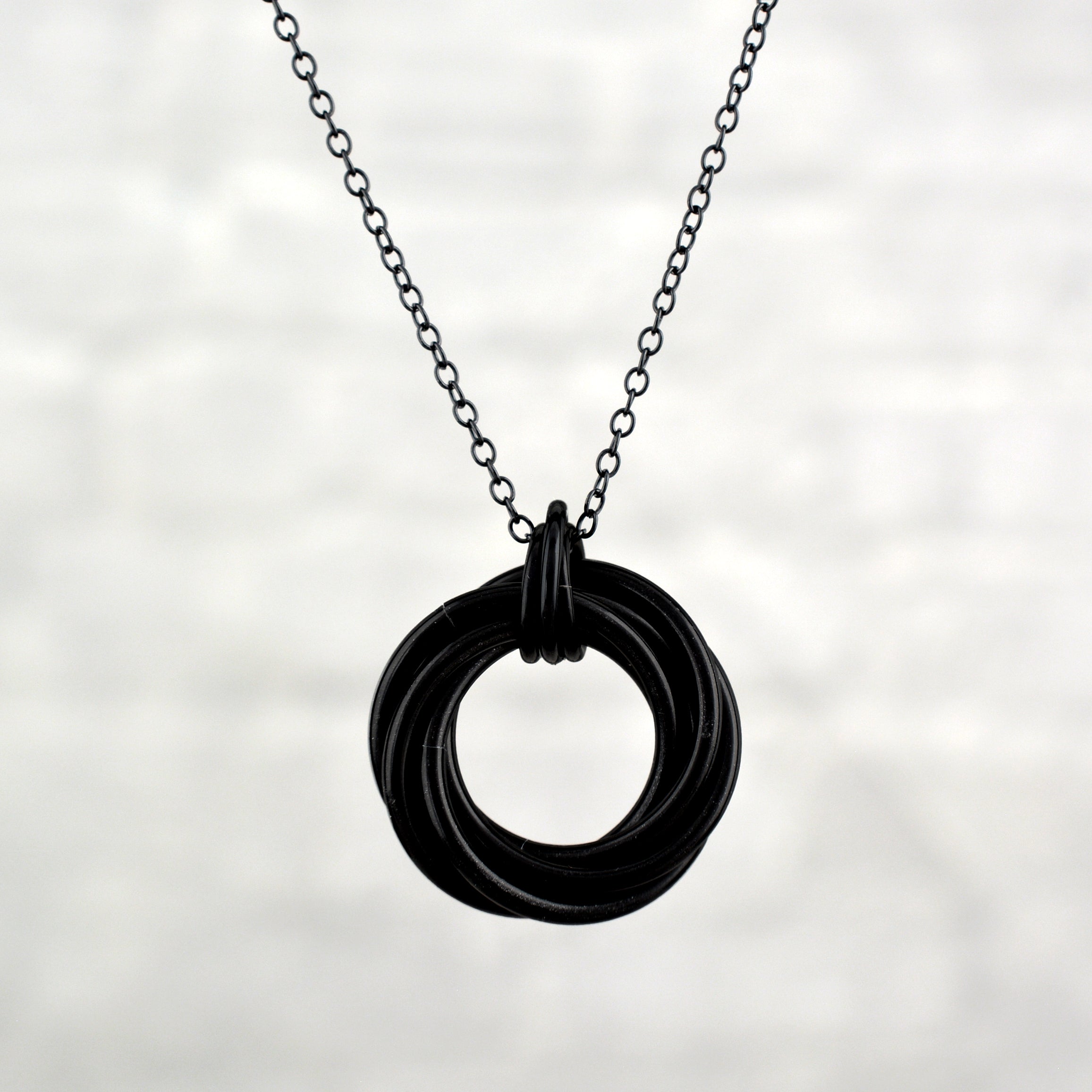 A circular black chainmaille pendant featuring intertwined rings, joined in a swirl, hangs on a delicate black chain against a textured light gray background.