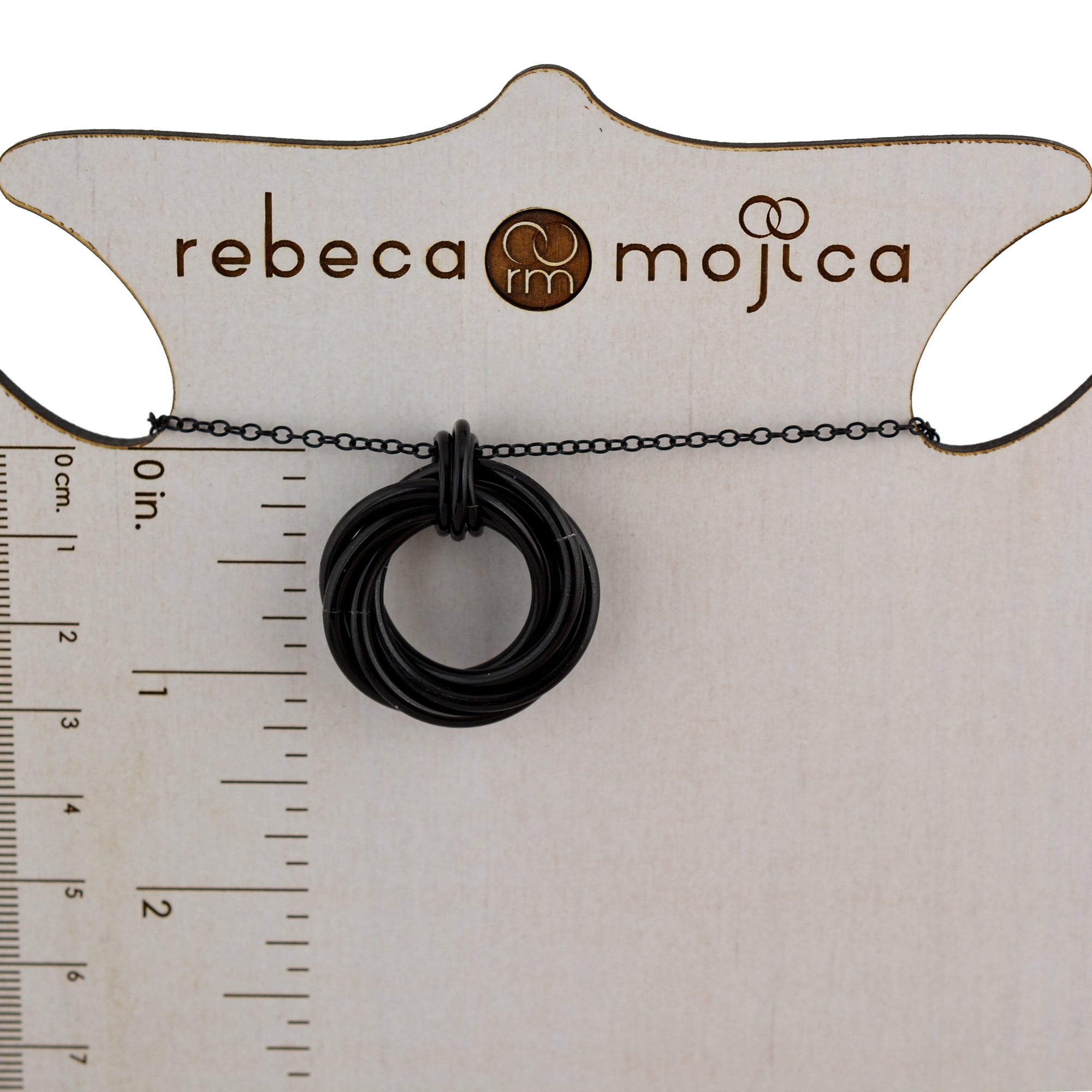 A black chainmaille swirl pendant on a black chain is displayed on a light-colored card with "rebeca mojica" branding. A ruler on the left edge indicates the pendant measures approximately 1.25 inches (32 mm).