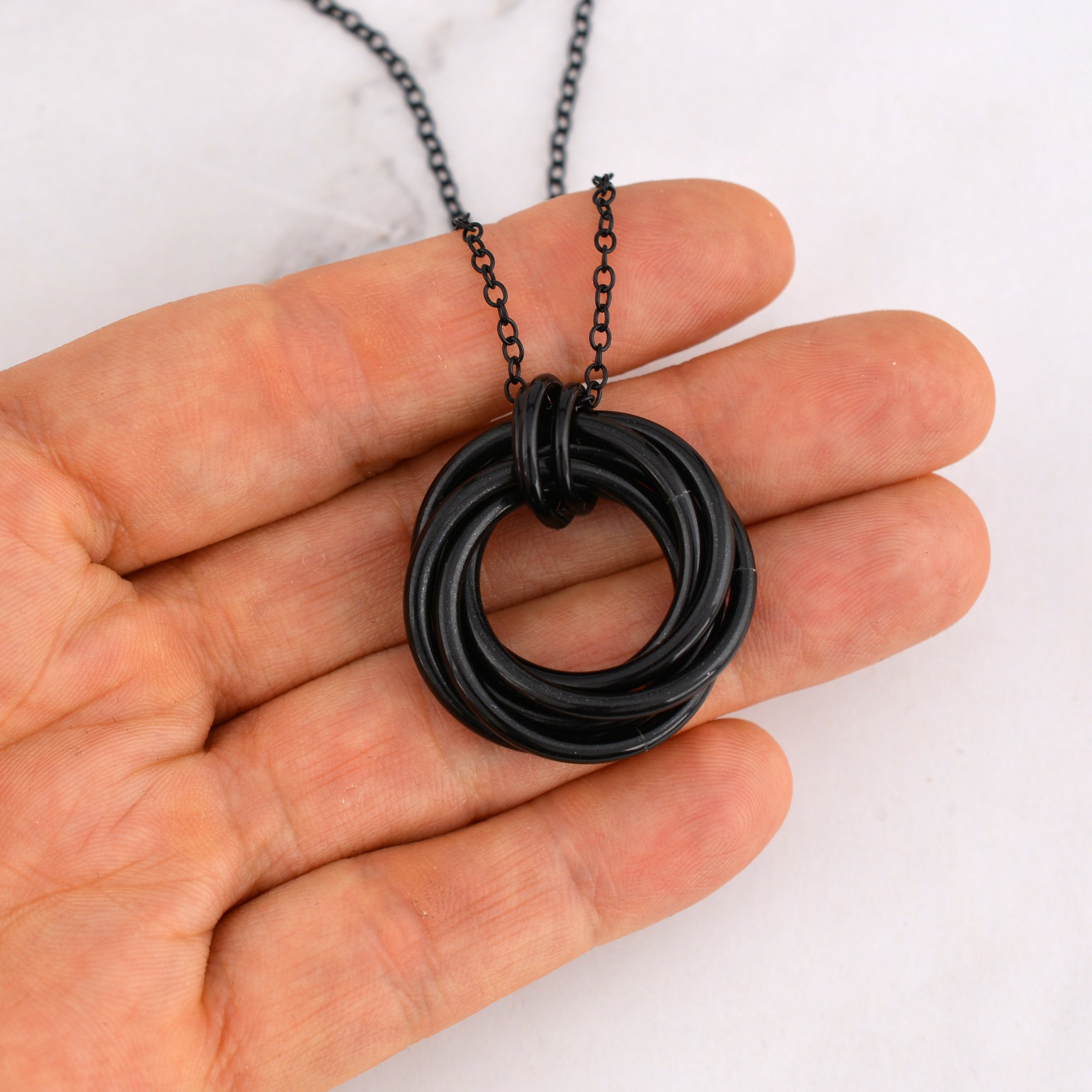 he black chainmaille swirl pendant rests on artisan Rebeca Mojica’s fingertips. The pendant hangs from a black chain, showcasing its size and highlighting its tactile nature against a light, marbled surface.