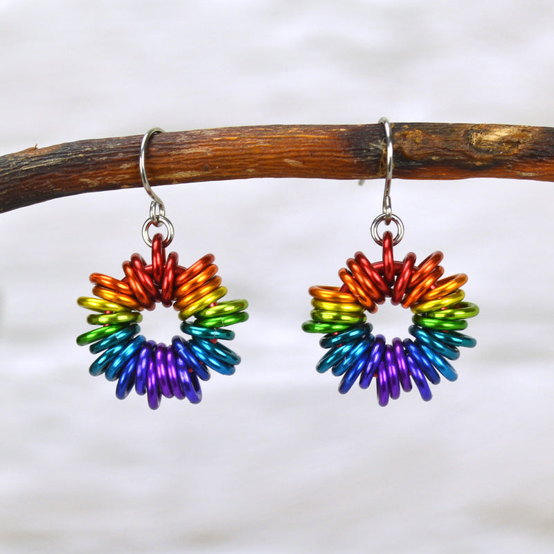 Rainbow Ombre Coiled Earrings by Rebeca Mojica Jewelry hanging from a branch. Each earring has 28 colorful jump rings added to a single large ring; the overall look resembles coiled wire. Colors range from red at the top, through orange, yellow, green blue and purple at the bottom.