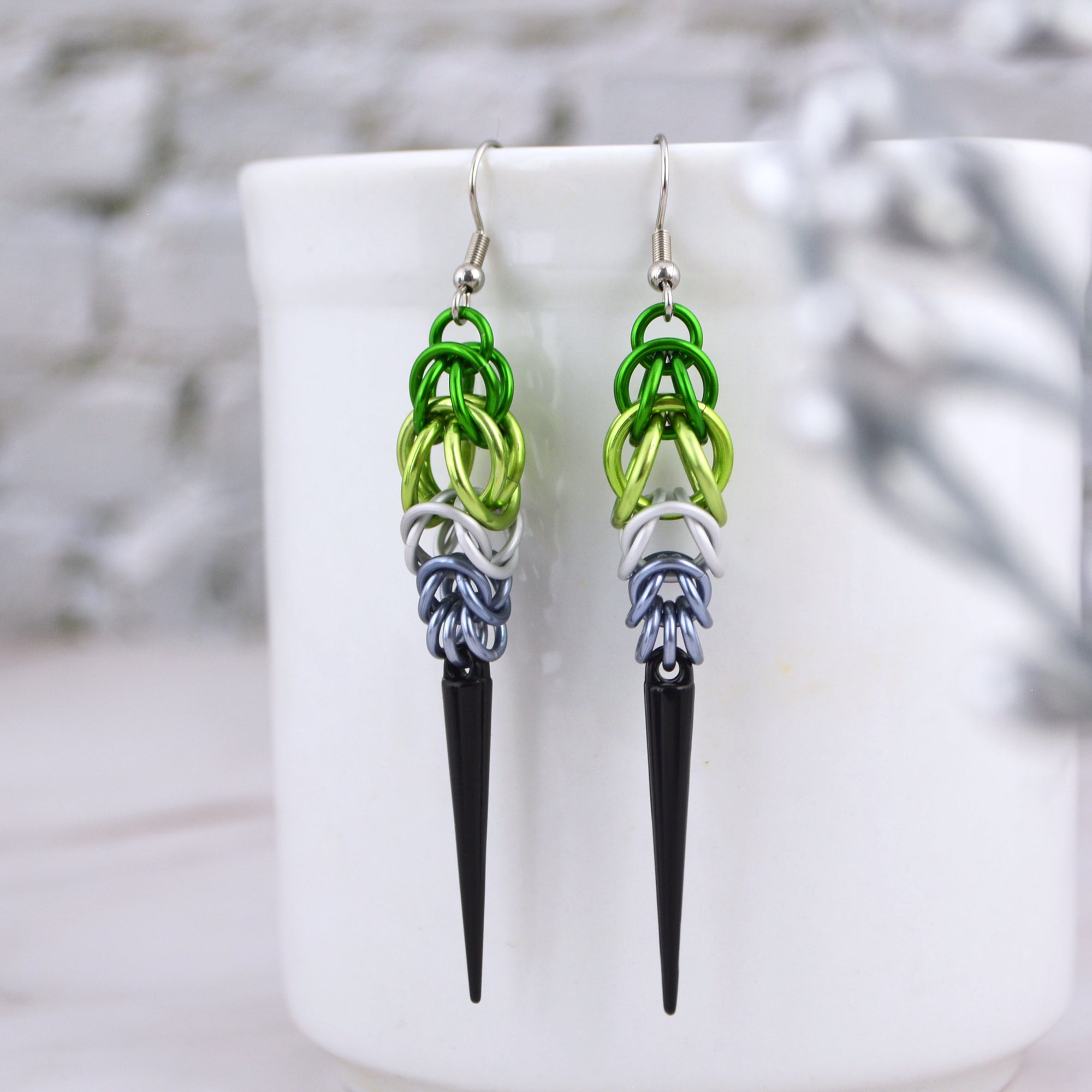Pair of aromantic pride chainmaille earrings hanging from a mug. The earrings have green rings at the top, white in the middle and grey at the bottom and are finished with a long black spike.