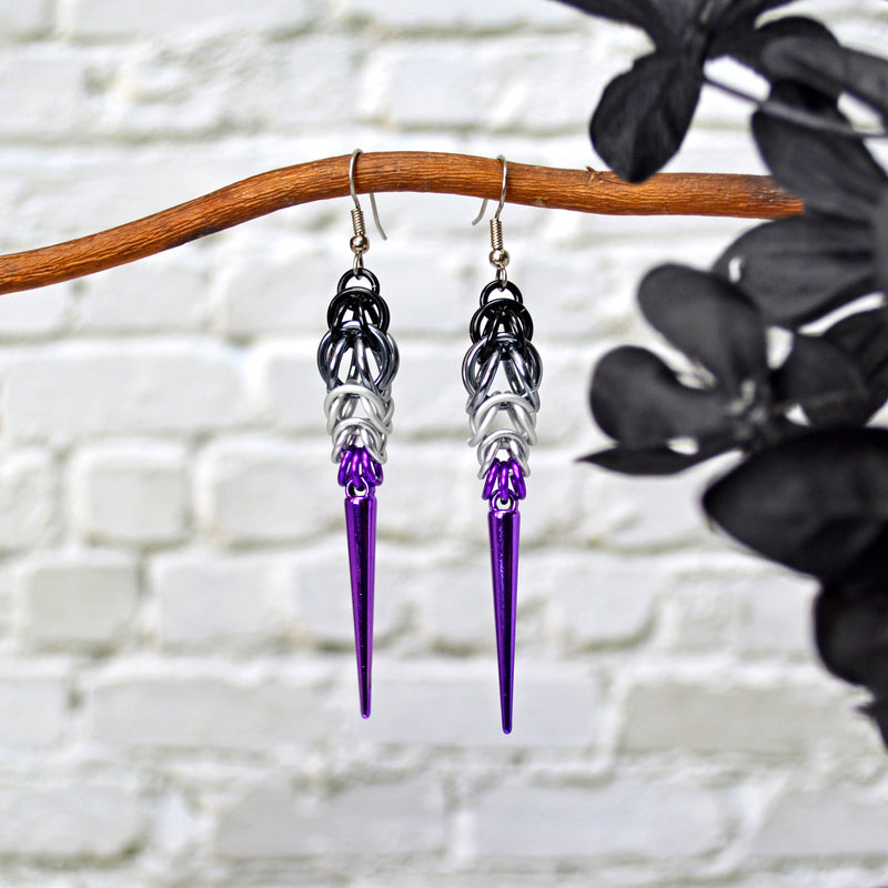 Asexual pride chainmaille earrings hanging from a branch. Earrings are black and grey at the top, followed by whitened violet with a dark lilac acrylic spike at the bottom.
