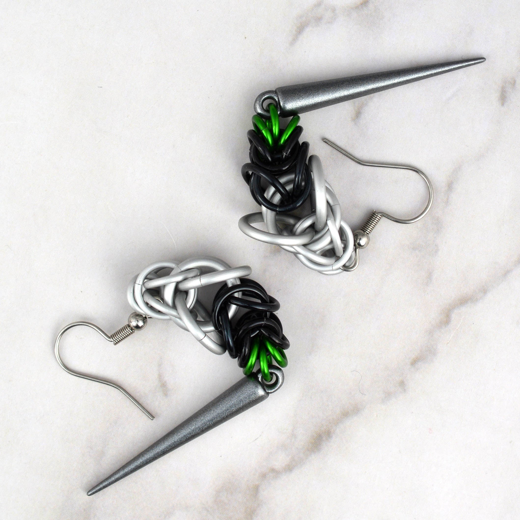A pair of Demiromantic Pride chainmaille earrings, handmade by artisan Rebeca Mojica, being held in her open palm. The chainmaille features white, black, and green aluminum rings, with a dark grey acrylic spike.
