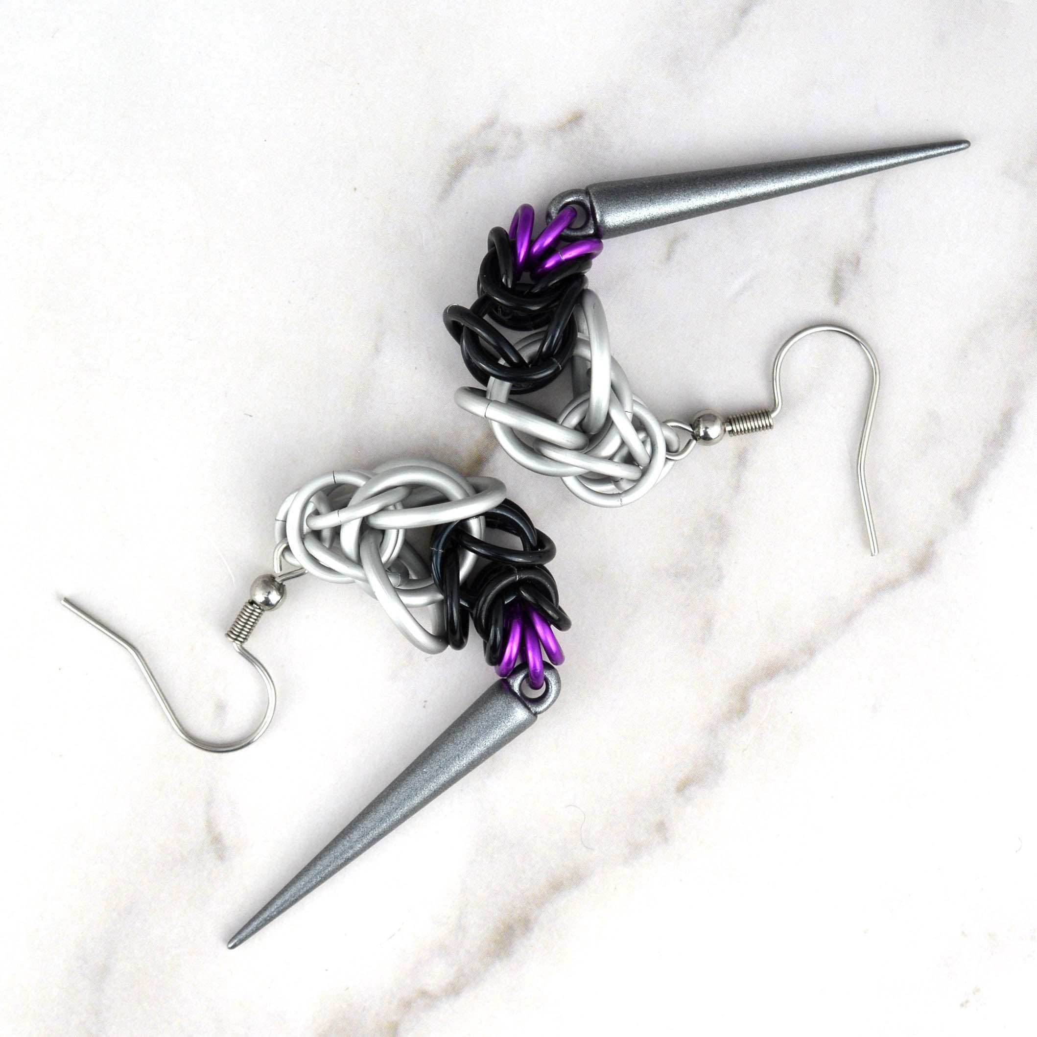 A pair of Demisexual Pride chainmaille earrings, handmade by artisan Rebeca Mojica, featuring white, black, and purple aluminum rings, with a long, dark grey acrylic spike dangling from each. The earrings are laid across a white marble surface, highlighting the flexibility of the chainmaille weave.