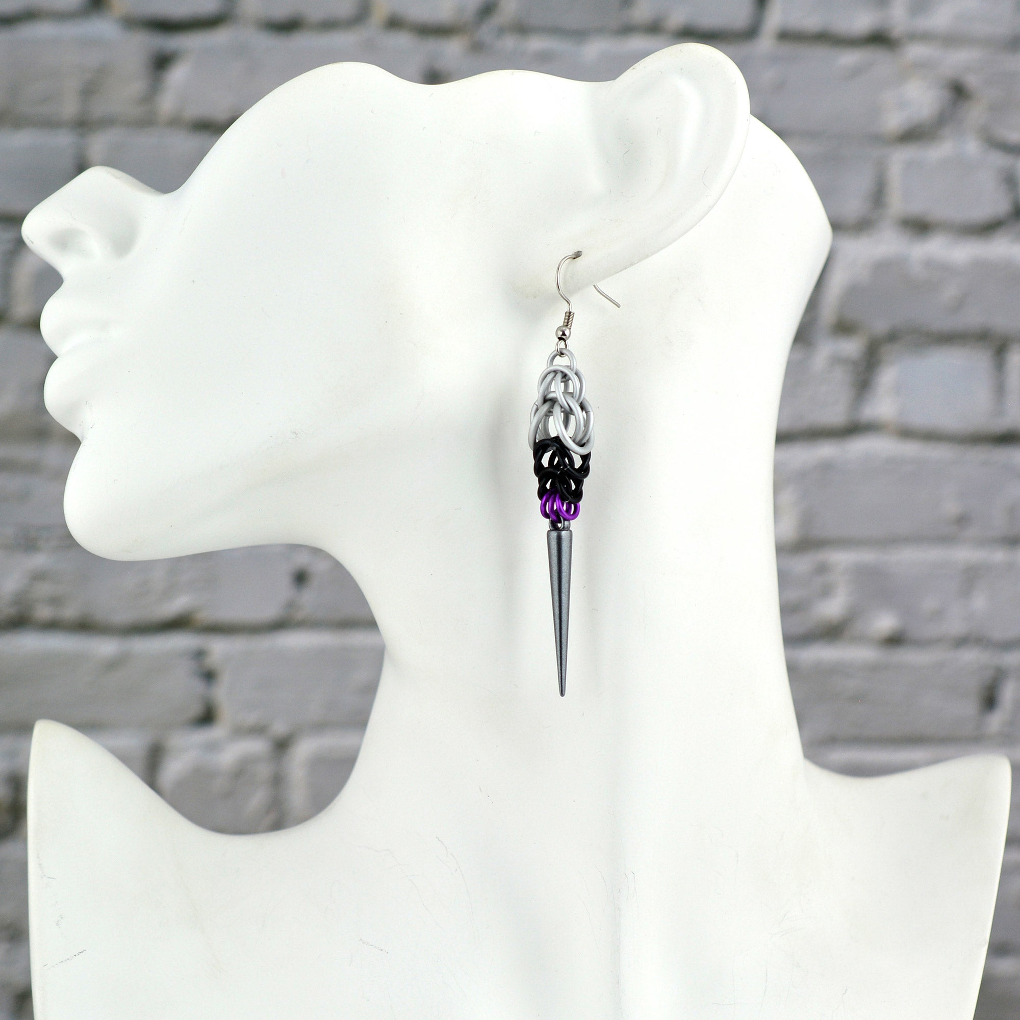 A Demisexual Pride spike earring, handmade by artisan Rebeca Mojica, displayed on a white mannequin head, seen in profile against a blurred gray brick background. The earring hangs from the ear, clearly showing the length and colors (white, black, and purple chainmaille) leading down to the dark grey acrylic spike.