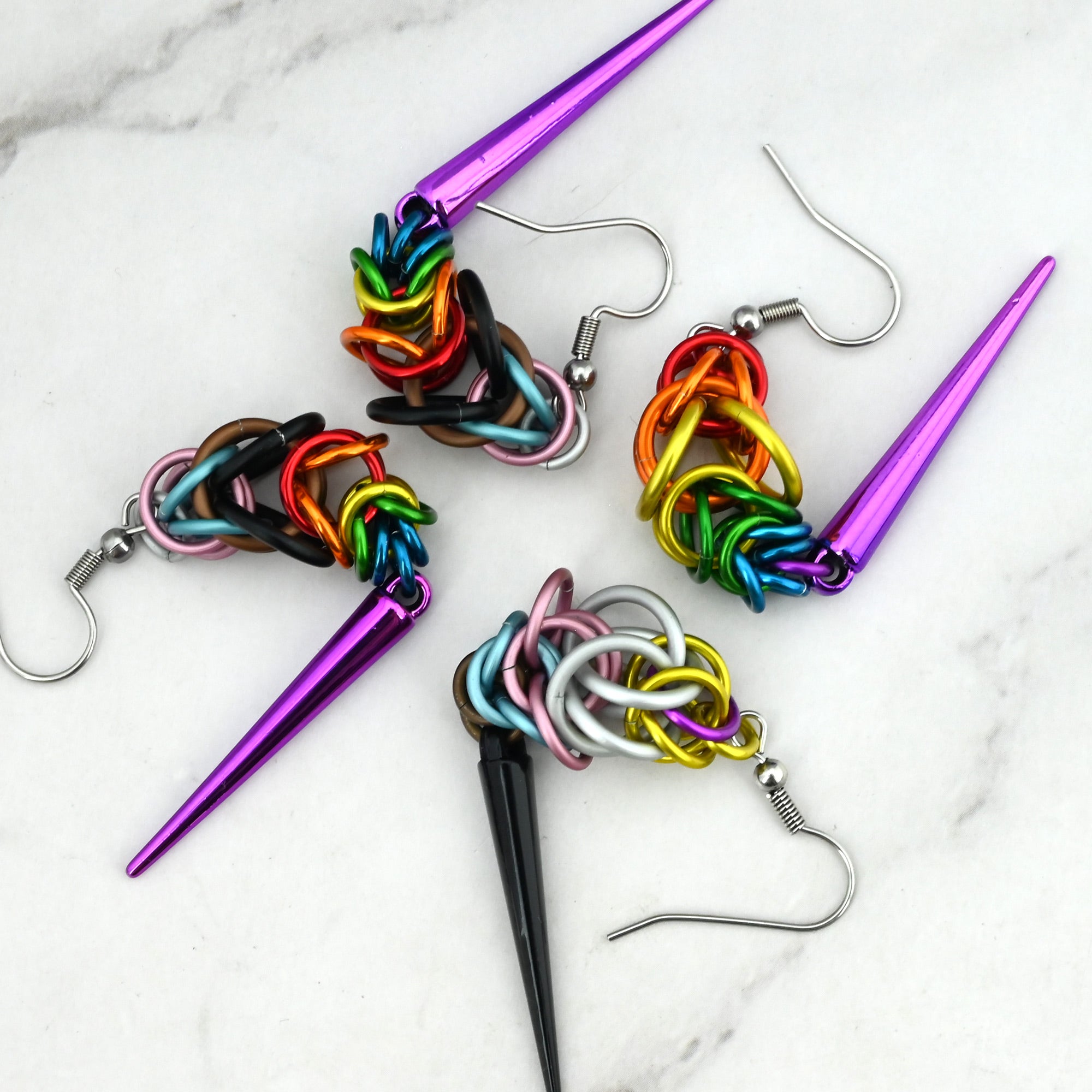 An overhead shot of four Progress Pride earrings on a marble surface. The earrings are curled and bent to demonstrate the flexible, fluid movement of the chainmaille weave.