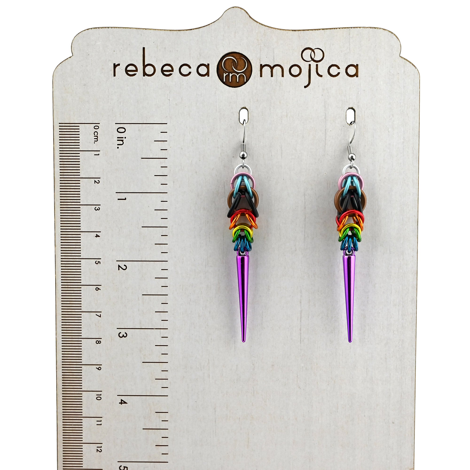 Two Progress Pride spike earrings hanging on a "Rebeca Mojica" display with a ruler, showing a total length of approximately 3.25 inches from the top of the earwire to the tip of the purple spike.