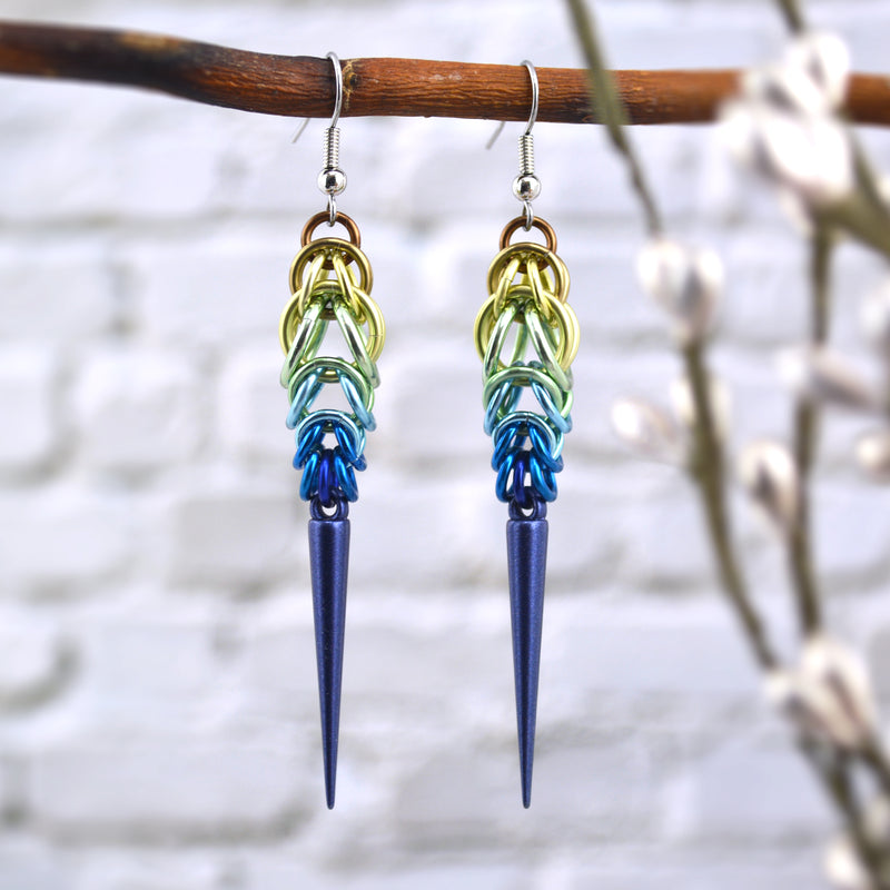 Chainmaille earrings in a striking brown-to-blue ombre hanging from a branch. Earrings are the Full Persian 6-in-1 weave and Box weave. Starting from the top, the colors are russet, champagne, pastel yellow, seafoam, light blue, azure and cobalt, with a cobalt spike. The background is a blurred off-white brick wall.