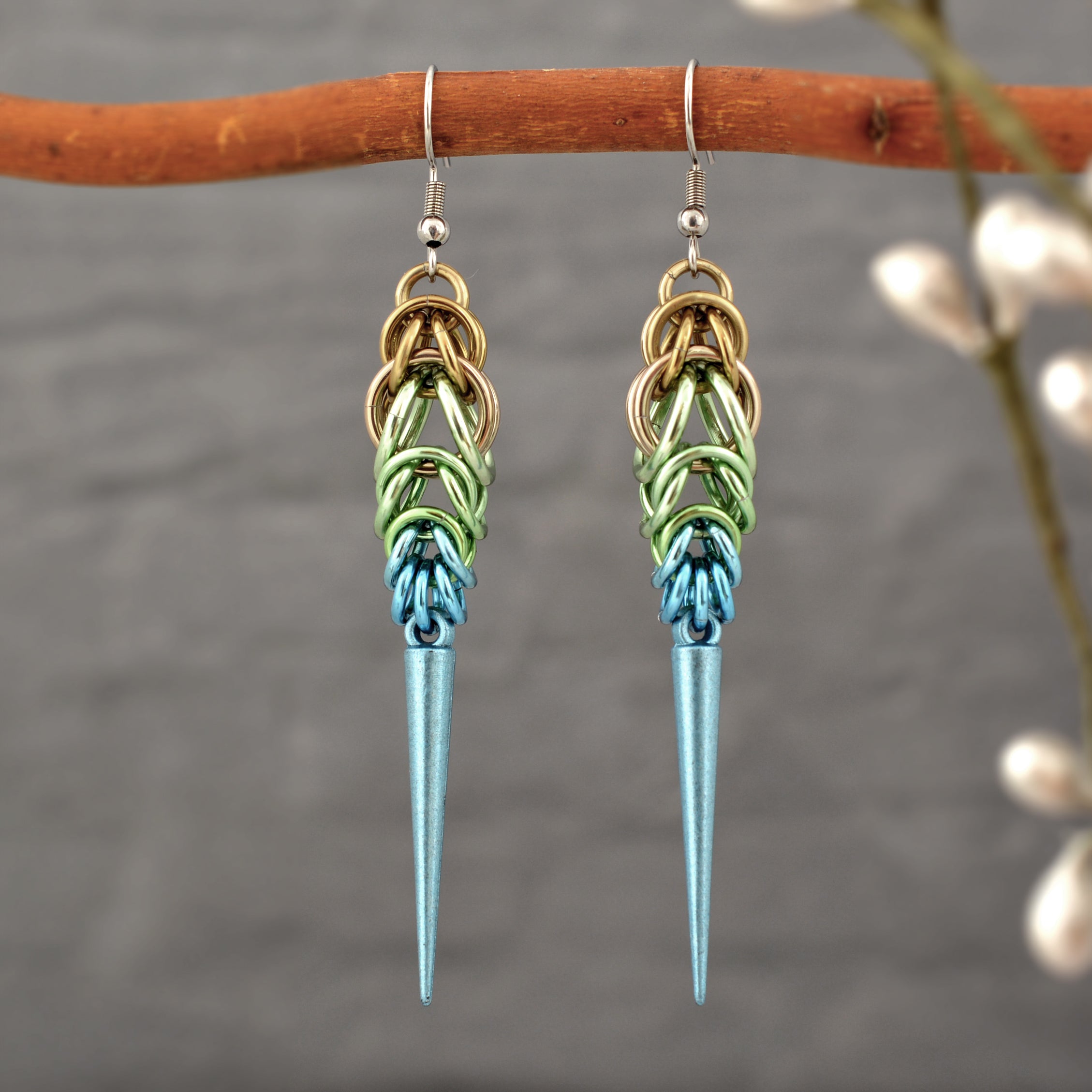 Handmade Seashell Ombre Chainmaille Spike Earrings hanging from a branch, with a soft-focus background of nature to emphasize their boho-edgy style. Earrings are a gradient of beige, seafoam and light blue chainmail links with a light blue spike at the base