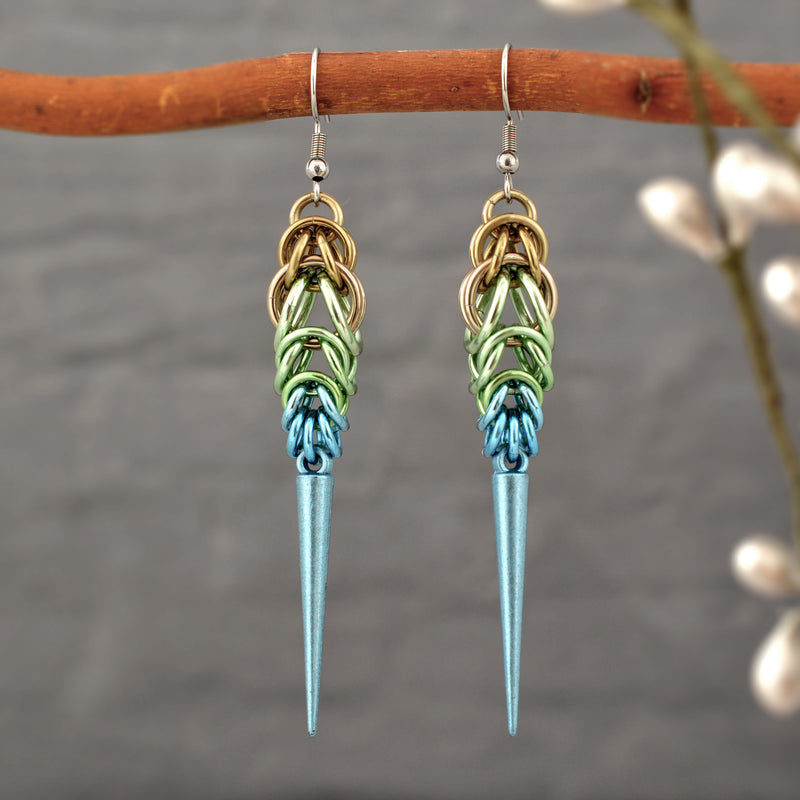 Handmade Seashell Ombre Chainmaille Spike Earrings hanging from a branch, with a soft-focus background of nature to emphasize their boho-edgy style. Earrings are a gradient of beige, seafoam and light blue chainmail links with a light blue spike at the base
