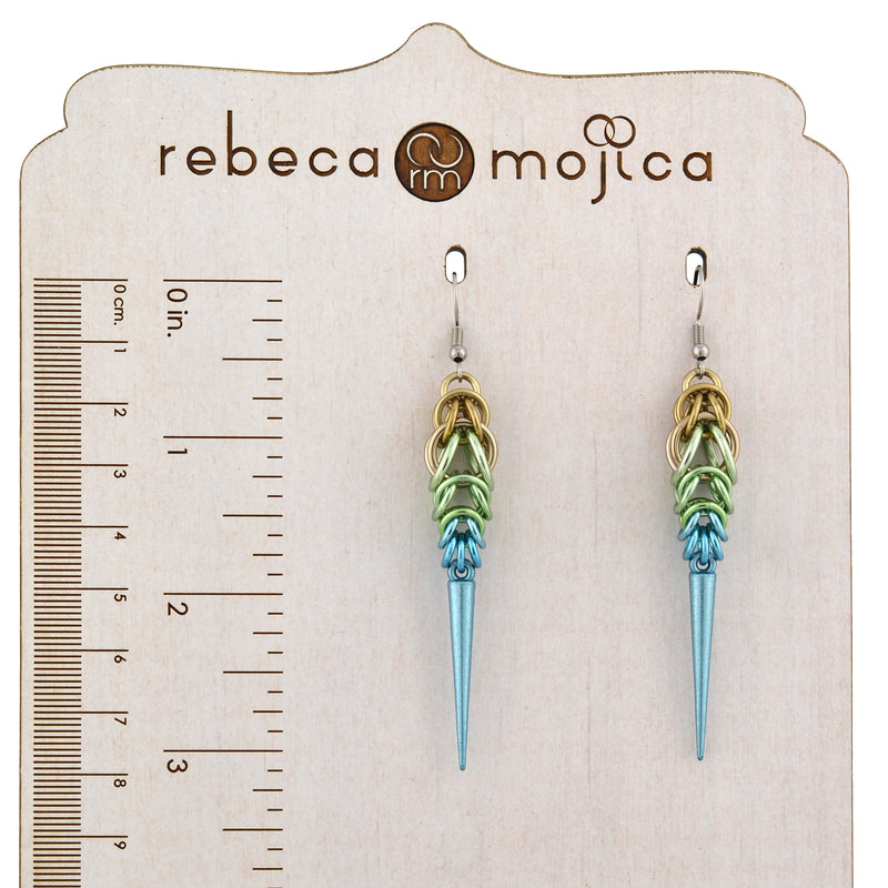 A pair of Rebeca Mojica Seashell Ombre Spike Earrings on a branded display board, with a ruler showing the length is about 3.25 inches.