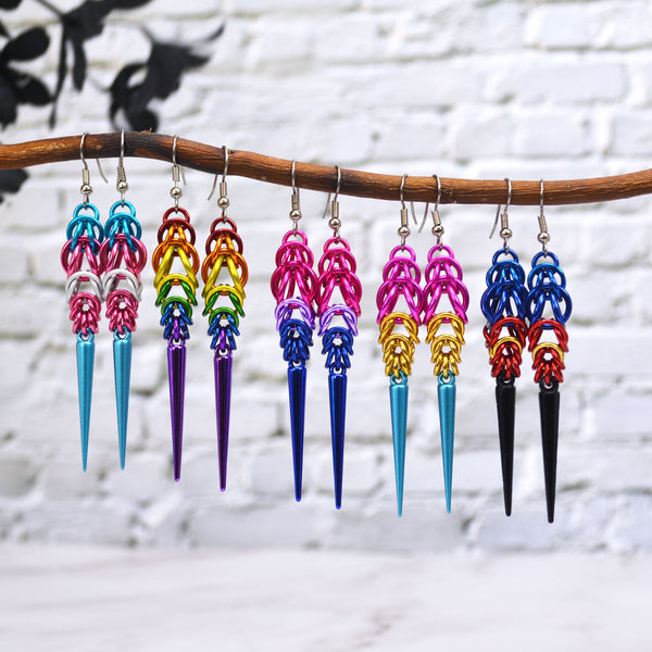 Colorful chainmaille earrings hanging from a branch against a white brick background. Each earring, made in the colors of a different pride flags, has chainmaille at the top in Full Persian and Box Weave, with a long acrylic spike at the base. The flags represented from left to right are: Transgender, Rainbow, Bisexual, Pansexual and Polyamory.