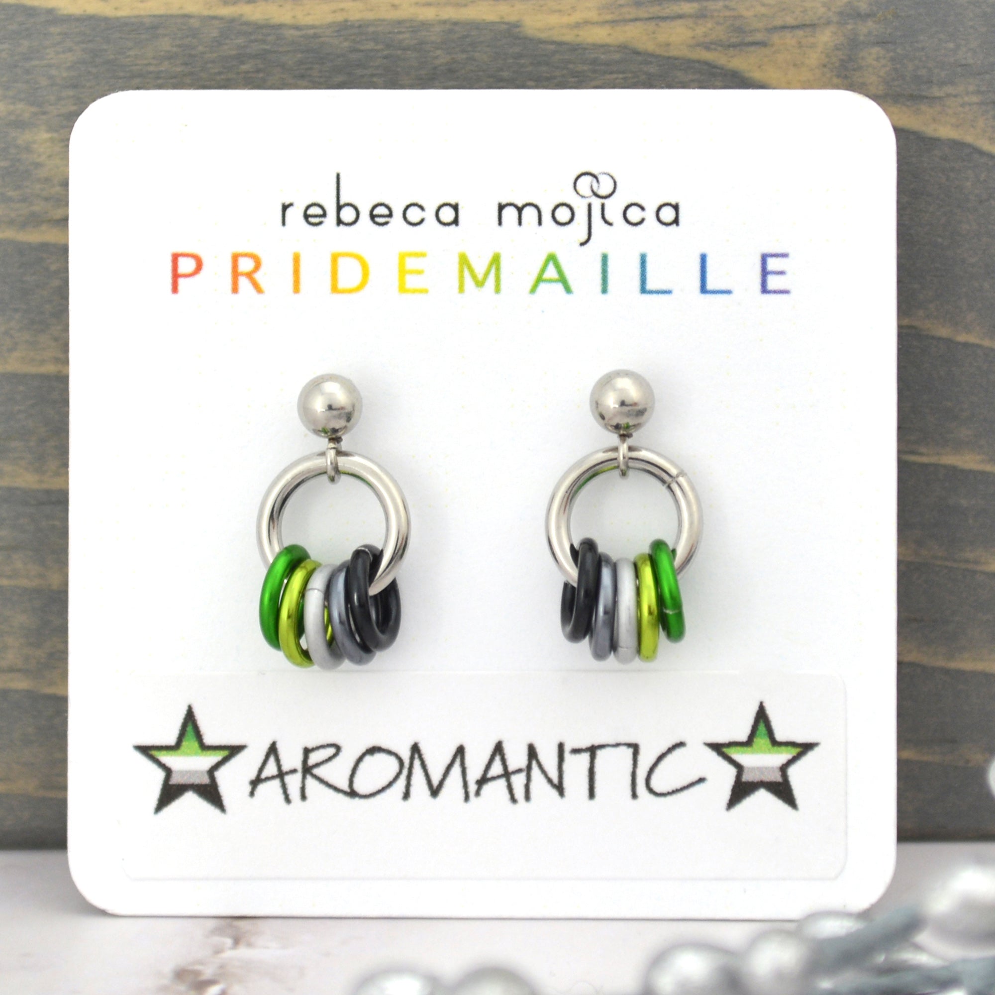Earrings on a card with 'rebeca mojica PRIDEMAILLE' branding and 'Aromantic' label. The earrings are small hoops attached to stud posts, with 5 tiny hanging links in the colors of the aromantic pride flag - green, light green, white, grey and black.