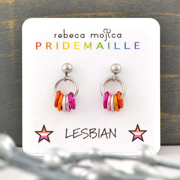 Tiny Lesbian Pride stud earrings displayed on a card. Earrings are on a post with a small steel ball. From the ball hangs a steel hoop that has 5 links hanging from it in the colors of the lesbian platonic pride flag: 2 shades of orange, white and 2 shades of pink. The earring card reads Rebeca Mojica at top, the second line reads PRIDEMAILLE in rainbow color fonts. The bottom of the card has a sticker with the word LESBIAN flanked by 2 stars in the color of the lesbian flag