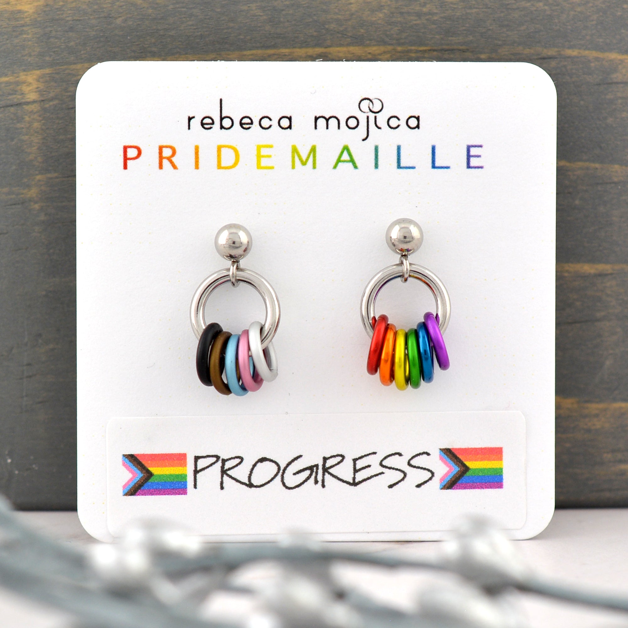A small pair of chainmaille earrings on a white card. The earrings are on posts with a small silver-color ball at top. Hanging from the ball is a steel jump ring, and attached onto that jump ring are small rings in the colors of the rainbow progress pride flag. The left earring has black, brown, light blue, pink and white. The right earring has red, orange, yellow, green, blue and purple. Card reads rebeca mojica PRIDEMAILLE progress
