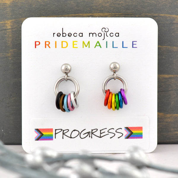 A small pair of chainmaille earrings on a white card. The earrings are on posts with a small silver-color ball at top. Hanging from the ball is a steel jump ring, and attached onto that jump ring are small rings in the colors of the rainbow progress pride flag. The left earring has black, brown, light blue, pink and white. The right earring has red, orange, yellow, green, blue and purple. Card reads rebeca mojica PRIDEMAILLE progress