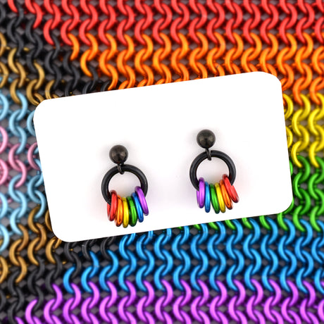 A pair of LGBTQIA+ pride stud earrings with black posts displayed on a white card. The background is a vibrant pattern of the European 4-in-1 chainmaille weave in rainbow-colored rings, creating the look of the Progress Pride Flag.