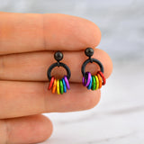 A pair of rainbow pride earrings held gently between chainmaille artisan Rebeca Mojica’s fingertips. The earrings feature black ball stud posts and a black ring, with six small rainbow-colored rings dangling below.