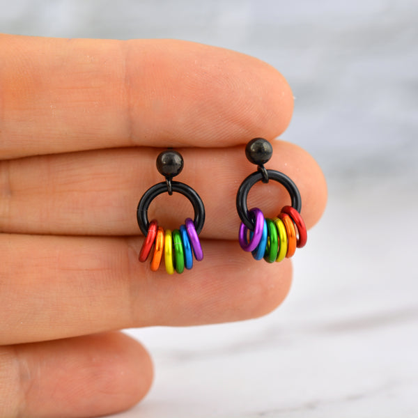 A pair of rainbow pride earrings held gently between chainmaille artisan Rebeca Mojica’s fingertips. The earrings feature black ball stud posts and a black ring, with six small rainbow-colored rings dangling below.