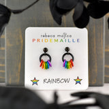 A pair of rainbow pride earrings resting on a white display card that reads "rebeca mojica PRIDEMAILLE" at the top and "RAINBOW" with two stars at the bottom. The card is standing against a dark gray wooden surface.