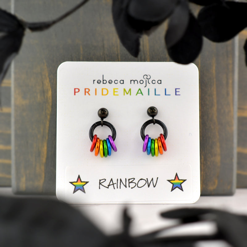 A pair of rainbow pride earrings resting on a white display card that reads "rebeca mojica PRIDEMAILLE" at the top and "RAINBOW" with two stars at the bottom. The card is standing against a dark gray wooden surface.