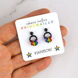 A small square white display card featuring two rainbow pride earrings is balanced on chainmaille artisan Rebeca Mojica’s fingertips. The card reads "rebeca mojica PRIDEMAILLE" and "RAINBOW" with stars.