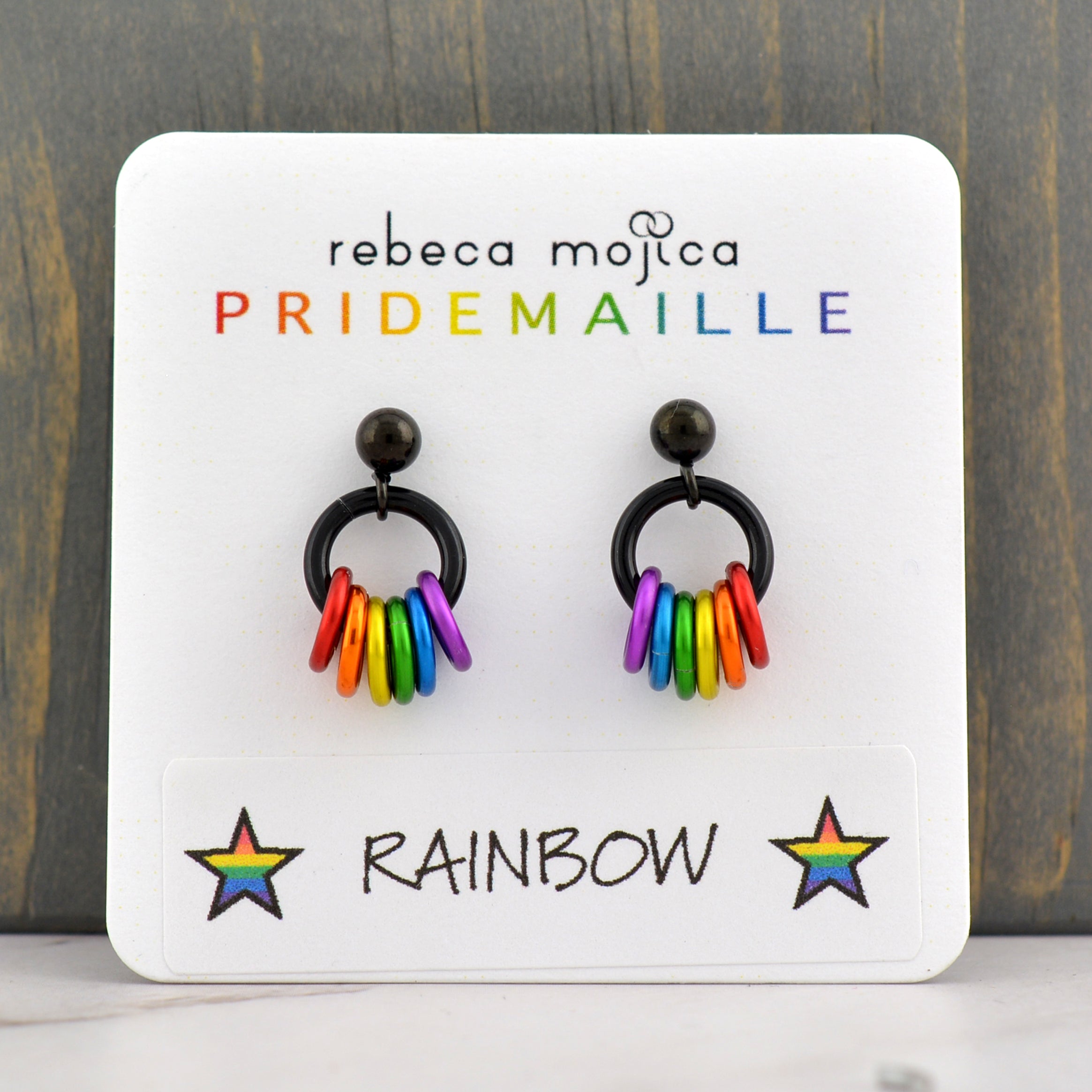 A pair of rainbow pride earrings on a white display card, showcasing the brand "rebeca mojica PRIDEMAILLE" and the "RAINBOW" color name with stars. The card is set against a dark wooden background.