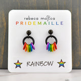 A pair of rainbow pride earrings on a white display card, showcasing the brand "rebeca mojica PRIDEMAILLE" and the "RAINBOW" color name with stars. The card is set against a dark wooden background.