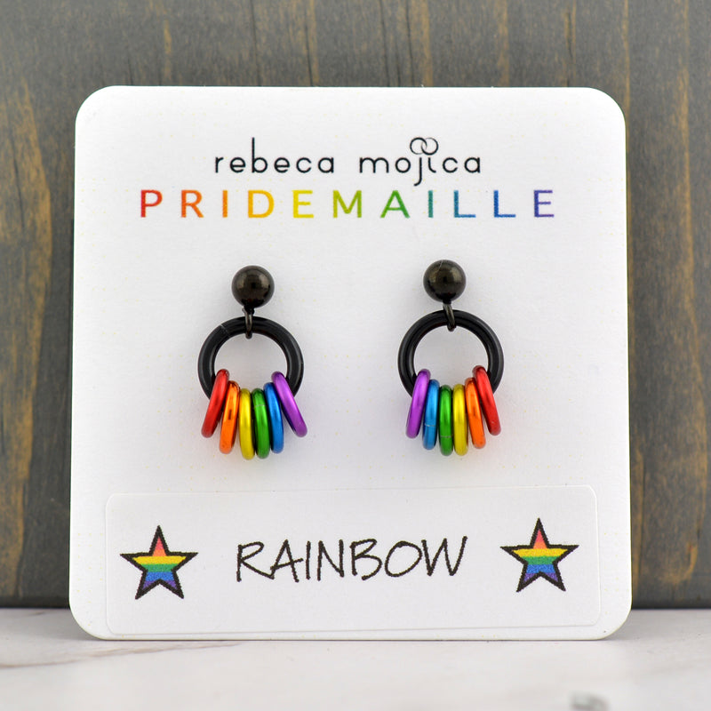 A pair of rainbow pride earrings on a white display card, showcasing the brand "rebeca mojica PRIDEMAILLE" and the "RAINBOW" color name with stars. The card is set against a dark wooden background.