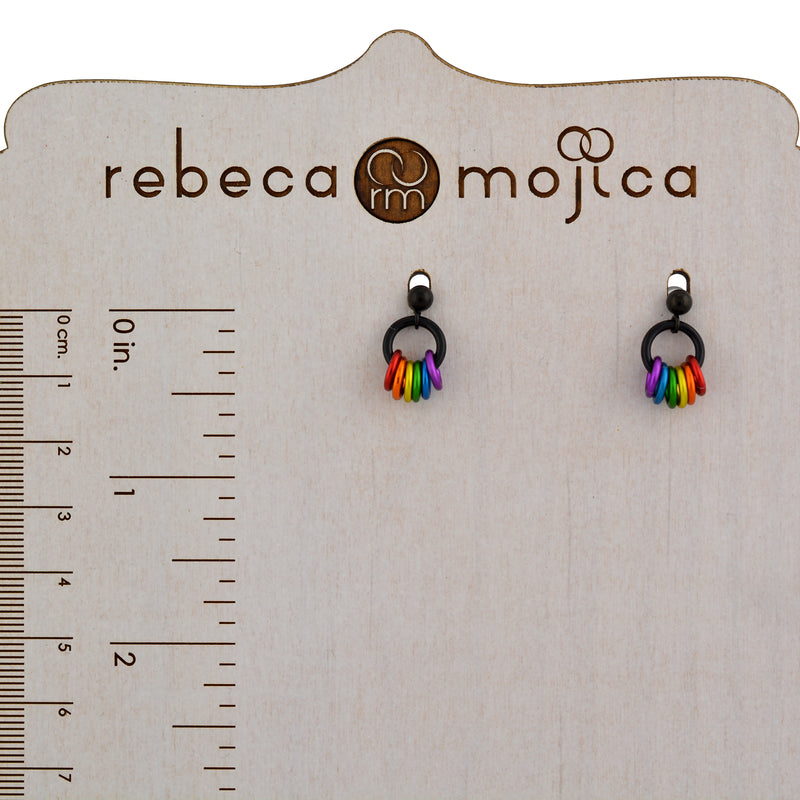 A pair of black post rainbow pride earrings displayed on a light wooden stand with a ruler measurement on the left side, showing dimensions in both centimeters and inches. These earrings measure about 0.5” from the top of the post to the bottom of the colorful links. The brand "rebeca mojica" is visible at the top.