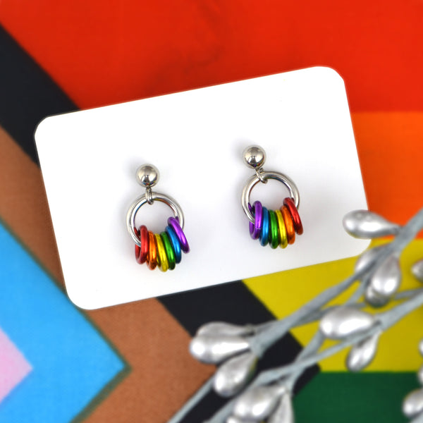 A small pair of chainmaille earrings on a white, resting against progress pride flag background. The earrings are on posts with a small silver-color ball at top. Hanging from the ball is a steel jump ring, and attached onto that jump ring are small rings in the colors of the rainbow pride flag: red, orange, yellow, green, blue and purple. Each earring is a mirror image of the other. Handcrafted rainbow pride chainmaille earrings by artisan Rebeca Mojica