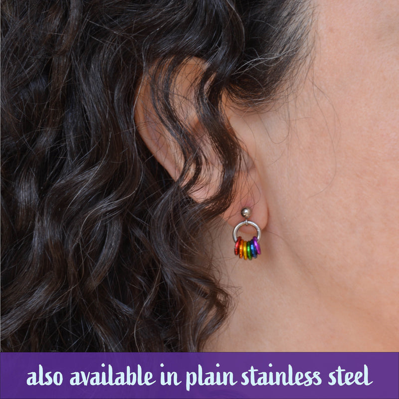 Close-up of Rebeca Mojica’s ear wearing a silver stud earring with small rainbow-colored rings dangling, highlighting the dainty size of the earrings. The image also displays the text "also available in plain stainless steel" at the bottom.
