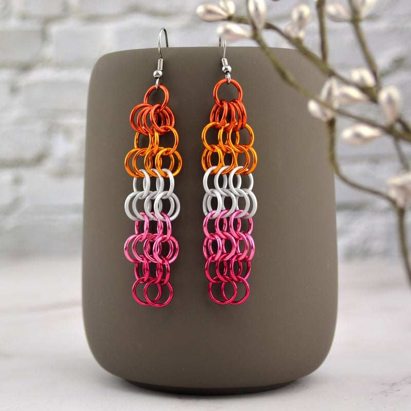A pair of 3-inch-long chainmaille mesh earrings hanging from the top of a grey mug. The earrings are rectangular shape with alternating rows of 3 and 4 links. There are two rows of each color of the lesbian pride flag, from top to bottom: dark orange, bright orange, white, pink and fuchsia. Background is an off-white brick wall and marble floor.
