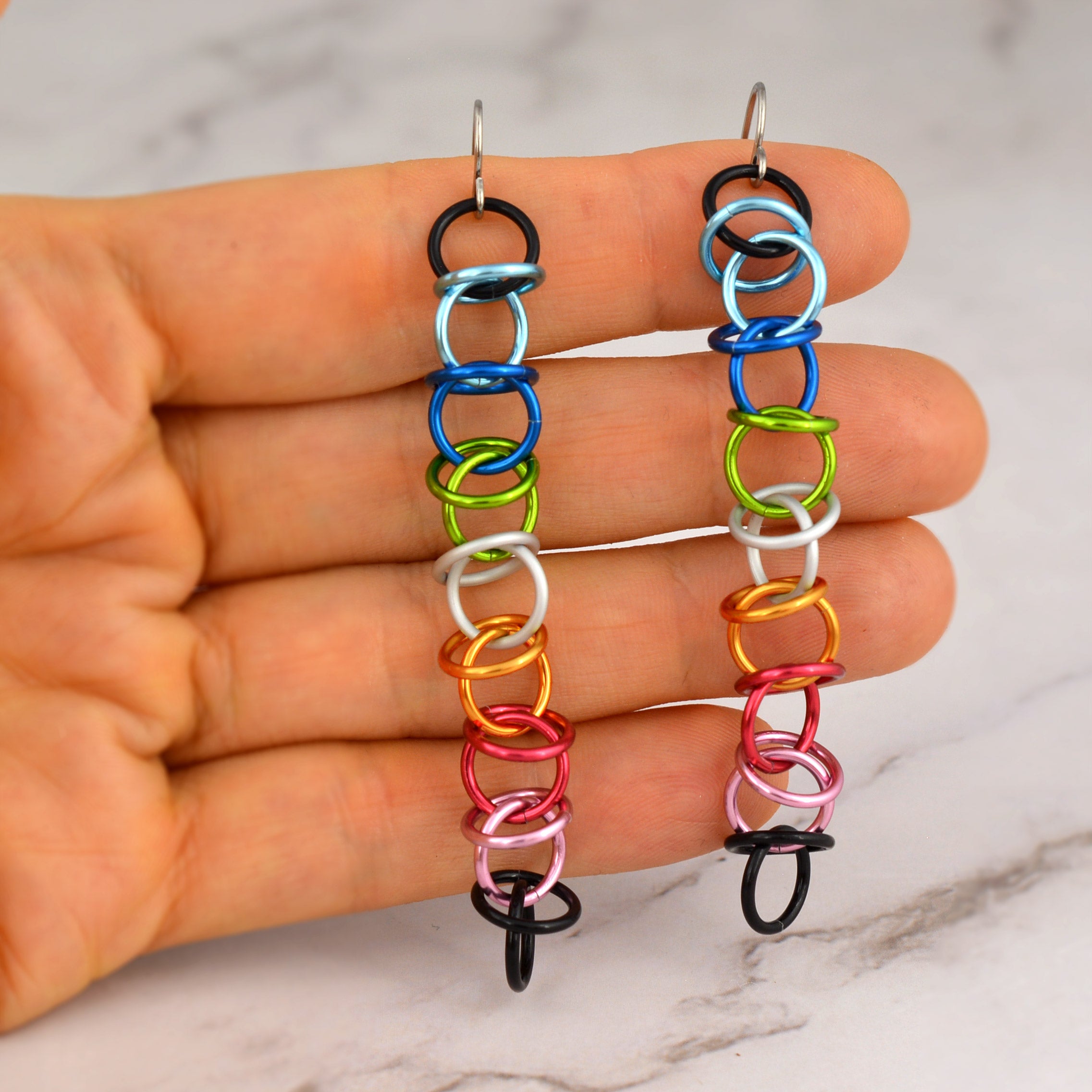 A close-up features long, dangling earrings held in the hand of chainmaille artist Rebeca Mojica against a softly blurred marble backdrop. The earrings showcase small, vibrant aluminum rings linked in an orbital chainmaille pattern. The colors represent the Queer Pride flag: black, light blue, blue, bright green, white, gold, dark pink, pink, and black links at the base. They are finished with simple stainless steel earwires.