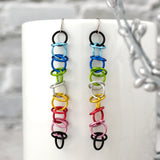 A pair of Queer Pride orbital earrings are showcased against a blurred setting of a smooth white mug and a touch of silver foliage. The earrings are composed of interconnected, vibrant aluminum rings in the Queer Pride flag colors: black, light blue, blue, bright green, white, gold, dark pink, pink, and black. Simple surgical steel earwires complete the design, highlighting their elegant dangle and vibrant colors.