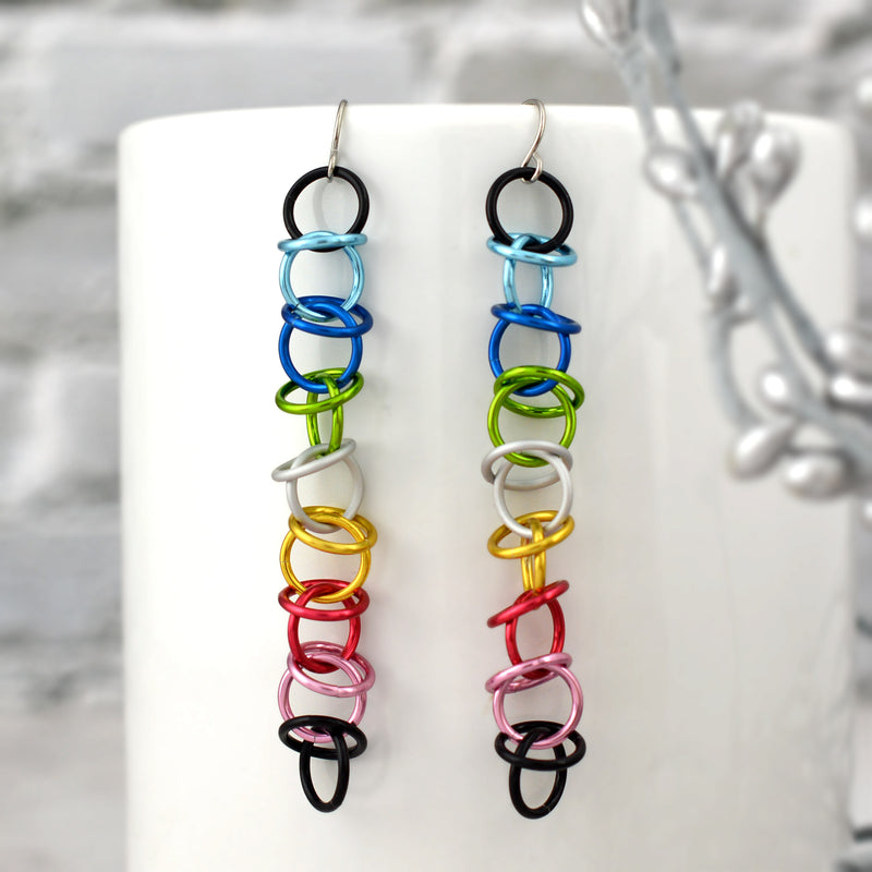 A pair of Queer Pride orbital earrings are showcased against a blurred setting of a smooth white mug and a touch of silver foliage. The earrings are composed of interconnected, vibrant aluminum rings in the Queer Pride flag colors: black, light blue, blue, bright green, white, gold, dark pink, pink, and black. Simple surgical steel earwires complete the design, highlighting their elegant dangle and vibrant colors.