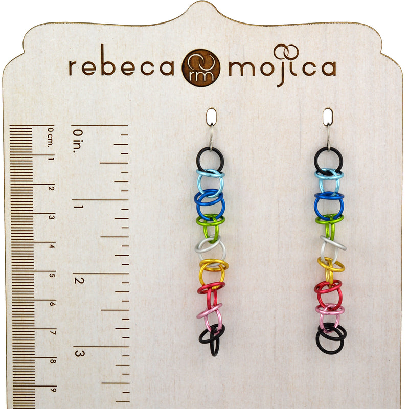 A pair of Queer Pride orbital earrings are displayed on a branded wooden stand, which includes a ruler marked in both centimeters and inches. The earrings are crafted from small, colorful aluminum rings linked together to show the Queer Pride flag colors. The ruler indicates an approximate length of 3.25 inches, and the stand features the "rebeca mojica" logo at the top.