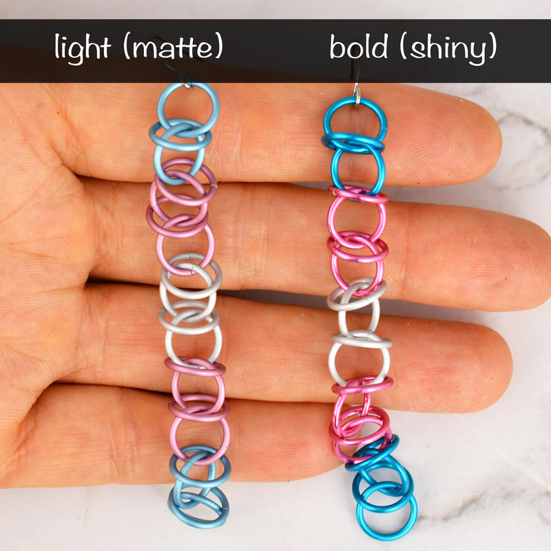 Transgender Pride Earrings Comparison: Two long, lightweight chainmaille earrings on Rebeca Mojica's hand. Left: light matte trans pride flag colors (light blue, pink, white). Right: bold shiny trans pride flag colors.