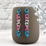Shiny Transgender Pride Earrings on Mug: Long, lightweight chainmaille earrings (bright trans pride flag colors) hanging from a grey mug. Steel earwires, blurred silver leaf background.