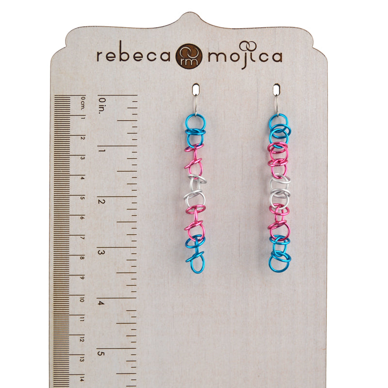 Trans Pride Earrings Length: Long, lightweight chainmaille earrings (matte trans pride flag colors) on a branded Rebeca Mojica wooden stand. Approximately 3.5 inches long.