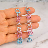 Handmade Transgender Pride Earrings: Close-up of long, lightweight chainmaille earrings in light blue, pink, and white on artisan Rebeca Mojica's hand. Surgical steel earwires, marble background.