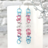Matte Transgender Pride Earrings on Mug: Long, lightweight chainmail earrings (light blue, pink, white) hanging from a white mug. Minimalist stainless steel earwires, blurred silver leaf background.