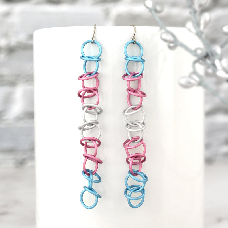 Matte Transgender Pride Earrings on Mug: Long, lightweight chainmail earrings (light blue, pink, white) hanging from a white mug. Minimalist stainless steel earwires, blurred silver leaf background.