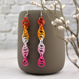 Long and thin chainmaille spiral earrings hang from a grey tumbler against a light grey brick wall and light grey marble surface. Earrings are the color of the lesbian pride flag: dark orange, bright orange, white, pink and fuchsia Earrings resemble a strand of DNA.